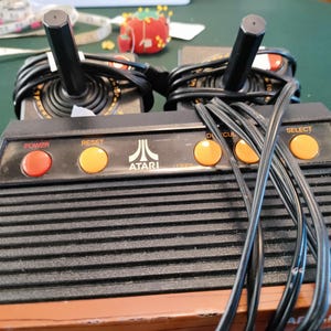 May include: A black Atari video game console featuring a ribbed top surface, a wood-grain base, and a control panel with a red power button, yellow reset, difficulty, and select buttons. Two black joysticks with coiled black cords are attached.