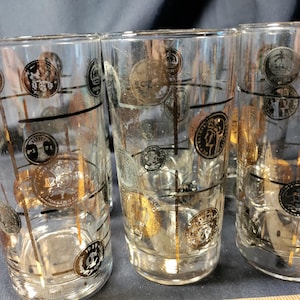 May include: Set of six vintage highball glasses. Each clear glass features gold and black coin designs and horizontal black lines. The glasses are cylindrical with a slightly flared base, perfect for serving cocktails or other beverages.