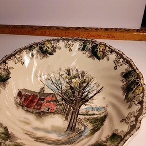 May include: A white ceramic bowl with a brown, green, and yellow floral pattern. The bowl features a scene of a red barn, a tree, and a stream. The bowl has a scalloped edge.