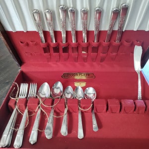 May include: A vintage silverware set in a wooden case with a red velvet interior. The set includes forks, spoons, knives, and a butter knife, all with ornate silver handles. The case has a Tudor Plate label.