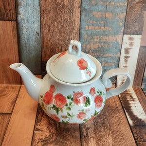 May include: White teapot with a floral design featuring pink roses and green leaves. The teapot has a lid and a handle.