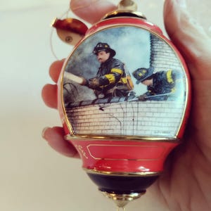 May include: Red and gold Christmas ornament with a detailed illustration of two firefighters on a rooftop. The ornament has a red base, a black bottom, and a gold-colored top. The illustration shows firefighters in action.