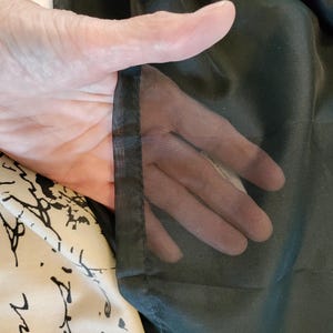 May include: A hand is holding up a sheer, black fabric, revealing the hand through the material. To the left is a cream-colored fabric with black script and abstract designs.