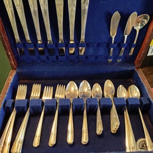 May include: A vintage silver-toned flatware set displayed in a wooden case with a blue velvet lining. The set includes forks, spoons, knives, and butter knives. The silverware has decorative handles and is arranged neatly within the case.