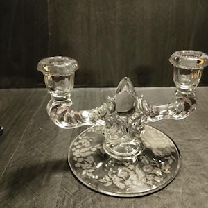 May include: A clear glass candle holder with a floral etched design. The holder has two candle cups and a twisted base.