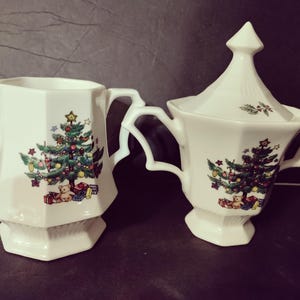 May include: White ceramic creamer and covered sugar bowl set. Each piece features a Christmas tree design with ornaments, gifts, and a teddy bear. The creamer has a spout and handle, while the sugar bowl has two handles and a conical lid.