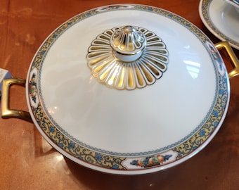 May include: A white porcelain serving dish with a gold and blue floral design. The dish has a lid with a gold knob and a gold floral design. The dish is on a wooden table.