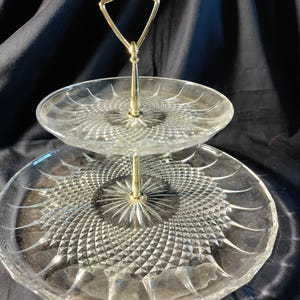 May include: A two-tiered glass serving tray with a gold-colored handle. The clear glass plates have a detailed, textured pattern. The handle is attached to the center of each plate, creating a decorative and functional piece for serving food or displaying items.