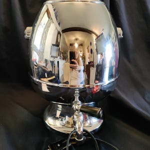 May include: A vintage chrome coffee urn with a glass lid and a spigot. The urn has a rounded body and a pedestal base. The shiny surface reflects the surrounding environment. A black power cord extends from the base.
