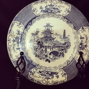 May include: A blue and white decorative plate with an Asian-inspired landscape design. The plate features a central scene of a pagoda, bridge, and floral elements, surrounded by a patterned border. The plate is displayed on a black metal stand.