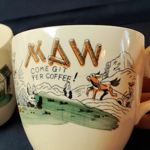 May include: A cream-colored ceramic mug featuring a graphic illustration of a horse-drawn cart in a landscape. The text "MAW" is written in orange log-style letters above the black text "COME GIT YER COFFEE!" with a green painted hill.