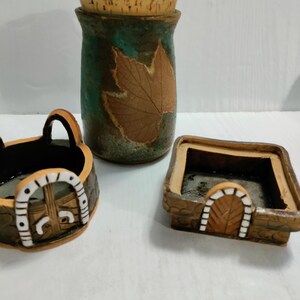 May include: Three ceramic containers with a cottagecore theme. The largest container is a jar with a cork lid and a brown leaf design. The other two containers are smaller and have a white archway design.