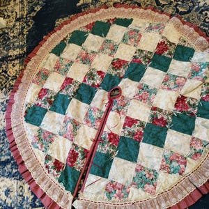 May include: A circular tree skirt featuring a patchwork design of alternating solid dark green squares and floral print squares in shades of red, pink, and white. The edge is finished with a ruffled dusty rose border and white lace trim.