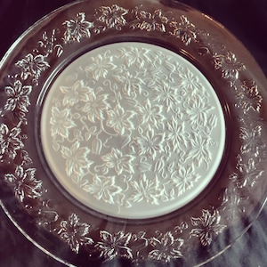 May include: Clear glass plate with a floral design. The plate has a raised floral pattern around the edge and a frosted floral design in the centre. The plate is round and appears to be made of glass.