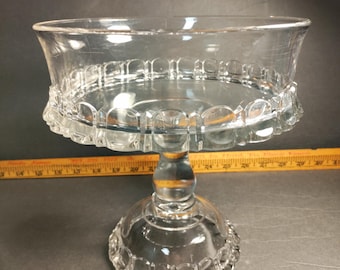 May include: A clear glass pedestal cake stand with a scalloped edge and a round base. The stand is about 7 inches in diameter.