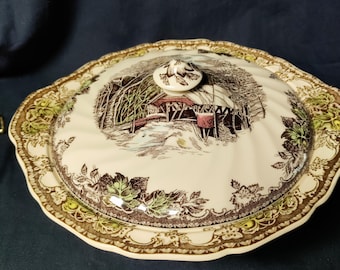 May include: A round ceramic serving dish with a lid, featuring a cream-colored base with a brown and green transferware pattern. The lid displays a detailed illustration of a covered bridge in a wooded landscape, topped with a small knob handle.
