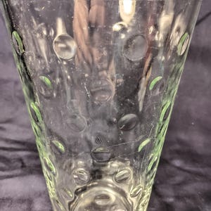 May include: A clear glass vase with a flared shape. The vase features a pattern of raised, circular dots. The glass has a slight green tint in some areas, and the vase appears to be empty.
