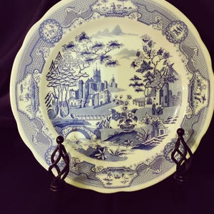 May include: A white and blue decorative plate with a landscape design. The plate features a detailed blue illustration of a village scene with buildings, trees, and a bridge. The plate has a scalloped edge and is displayed on a black stand.