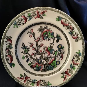 May include: A white ceramic plate with a floral design. The plate features a central image of a flowering tree with pink and red blossoms, surrounded by a black border and a decorative edge with floral motifs.