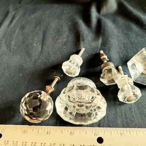 May include: A collection of clear glass cabinet knobs in various shapes, including faceted spheres, octagons, and rounded designs. The knobs have metal stems and are arranged on a dark surface. A ruler is in the foreground.