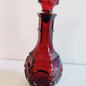 May include: A ruby red glass decanter with a textured stopper. The bottle has a geometric design with a diamond pattern and a faceted stopper. The decanter is approximately 10 inches tall.