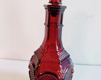 May include: A ruby red glass decanter with a textured stopper. The bottle has a geometric design with a diamond pattern and a faceted stopper. The decanter is approximately 10 inches tall.