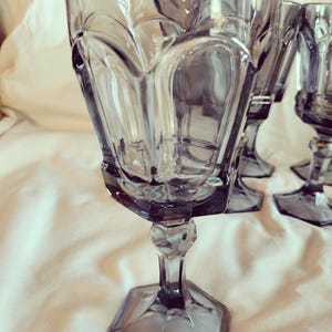 May include: Set of vintage gray-tinted drinking glasses. Each glass features a faceted base and stem, with a decorative pattern on the bowl. The glasses are arranged on a white surface, with soft lighting highlighting their details.