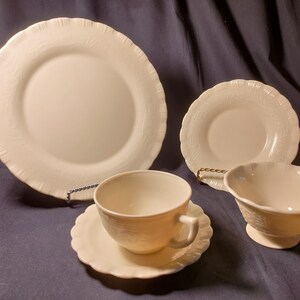 May include: A cream-colored, vintage dinnerware set. The set includes a large dinner plate, a smaller plate, a teacup with saucer, and a small bowl. All pieces have a textured, embossed design around the edges.