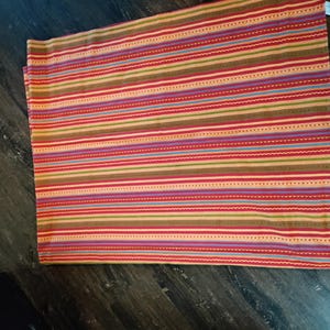 May include: A rectangular fabric textile featuring a repeating pattern of horizontal stripes in shades of red, orange, yellow, green, blue, and purple. The design includes thin solid lines, dotted lines, and wavy lines across the surface.