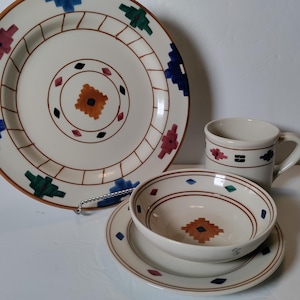 May include: A cream-colored ceramic dish set with a Southwestern design. The set includes a large plate, a bowl, a saucer, and a mug. The plates and bowl have a brown rim and feature geometric patterns in blue, green, red, and orange.