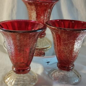 May include: Three red glass vases with a crackle finish and a flared top. The vases have a silver-colored base and a dark rim. The vases are arranged in a group, with one vase in the background and two in the foreground.