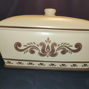 May include: Cream-colored ceramic bread box with a matching lid and a brown decorative floral pattern. The bread box has a rectangular shape with a slightly curved lid and a brown trim. The base features a row of brown heart-shaped designs.