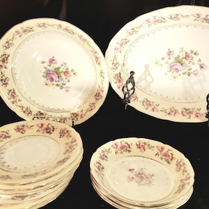May include: A collection of vintage porcelain dishes with a floral rose pattern. The set includes large serving bowls, plates, and smaller bowls. The edges are scalloped and trimmed with gold. The floral design features pink, purple, and yellow roses.