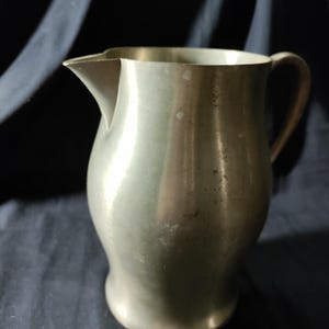 May include: A vintage metal pitcher with a curved body, spout, and handle. The pitcher has a silver-gray finish and shows signs of age. It is likely made of pewter or a similar metal. The design is simple and classic, suitable for serving beverages.