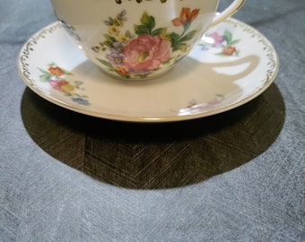 May include: A white teacup and saucer set with a floral pattern in shades of pink, yellow, blue, and green. The cup and saucer have a gold trim around the edges.