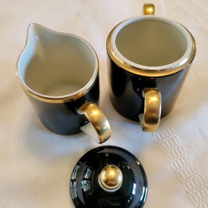 May include: A black and gold tea set, including a creamer, sugar bowl, and lid. The set features a glossy black finish with gold trim and handles. The creamer has a spout, and the sugar bowl has a lid with a gold knob. The set is on a white surface.