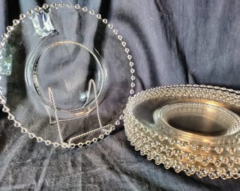 May include: Clear glass plates with beaded edges. One plate is displayed on a silver metal stand, while a stack of similar plates rests nearby. The plates have a circular design with a raised center.