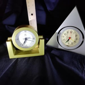 May include: Two decorative clocks. One is gold-colored, round, and sits on a rectangular base. The clock face has Roman numerals and the letters "PS". The other clock is triangular and silver, with a round clock face.