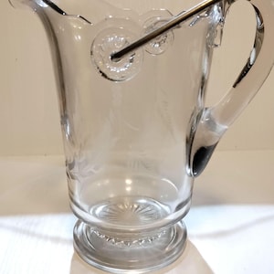 May include: A clear glass pitcher with a decorative handle and a glass stirrer. The pitcher has a scalloped rim and a round base.