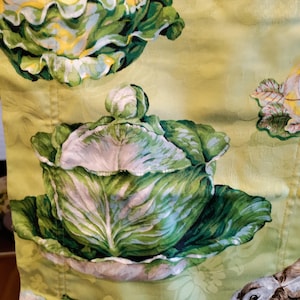 May include: A light green fabric featuring a repeating pattern of cabbage heads, including one shaped like a lidded tureen, alongside illustrations of rabbits and small green frogs. The textile has a subtle, tonal damask-style floral background.