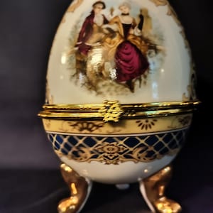 May include: An egg-shaped decorative trinket box with a hinged lid. The porcelain box features a painted scene of two figures in period clothing. Gold accents and feet support the box, with blue and gold patterned bands around the base.
