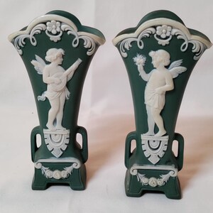May include: A pair of dark green vases featuring white relief carvings of winged figures. One figure plays a stringed instrument, while the other holds a bouquet. Both vases have ornate white floral and scrollwork detailing at the top and base.