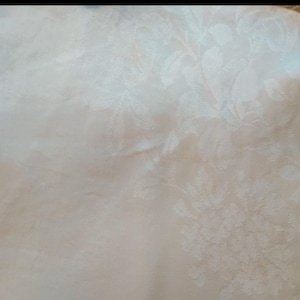May include: A white fabric featuring a tonal damask pattern with intricate floral and leaf motifs woven directly into the material. The surface displays a subtle sheen, highlighting the texture of the botanical design across the textile.