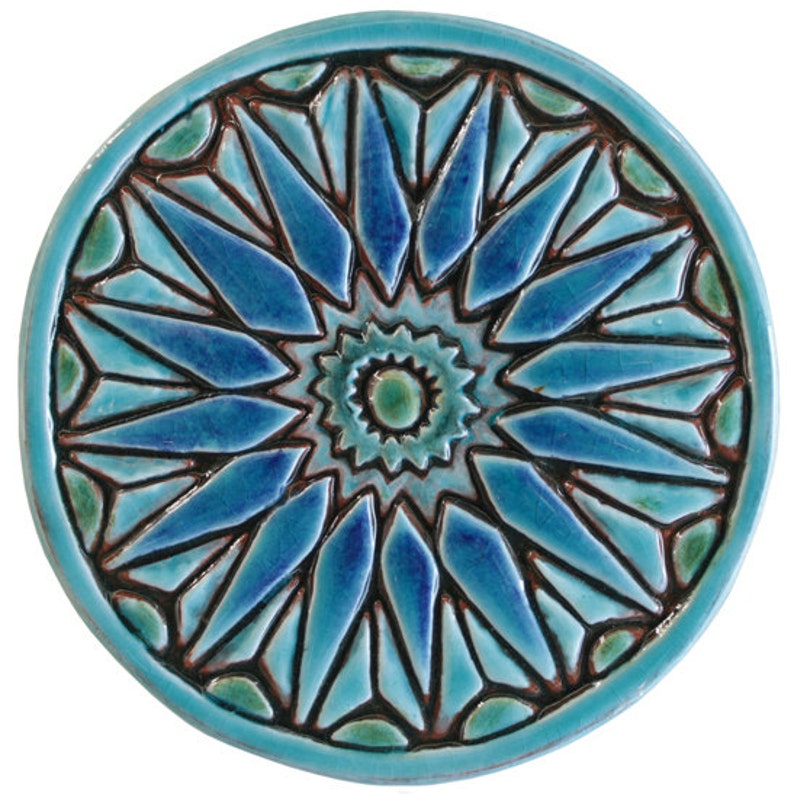 May include: A round ceramic tile with a blue and green geometric design. The design features a central starburst with radiating points.