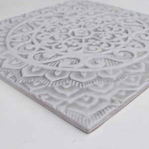 May include: A white ceramic tile with an intricate, embossed mandala design. The tile has a raised, textured surface and a square shape.
