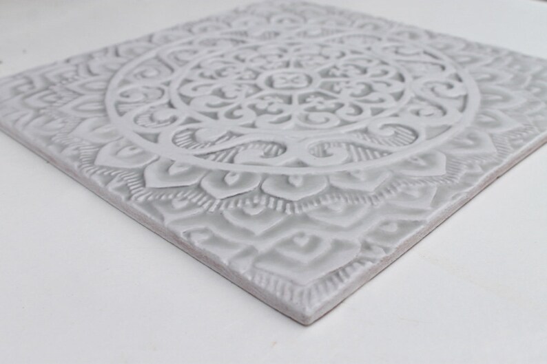 May include: A white ceramic tile with an intricate, embossed mandala design. The tile has a raised, textured surface and a square shape.
