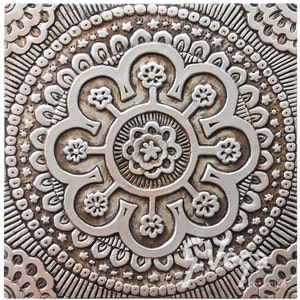 May include: A silver metal wall tile with a raised, embossed design of a circular mandala with a floral pattern. The tile has a distressed, aged look.