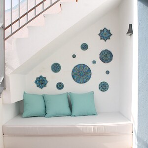 May include: A white couch with blue and green patterned throw pillows sits under a white staircase. The wall is decorated with blue and green patterned ceramic tiles in various shapes and sizes.