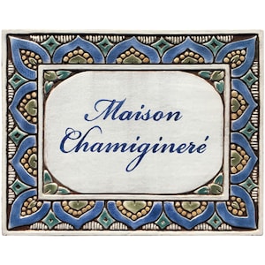 May include: A white ceramic tile with a blue and green border. The tile has the words "Maison Chamignere'" written in blue cursive script.