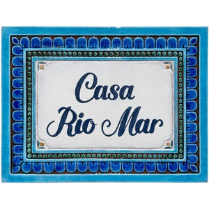 Op de afbeelding: Een blauwe en witte keramische tegel met een decoratieve rand. De tegel heeft de tekst "Casa Rio Mar" in een cursief schrift.